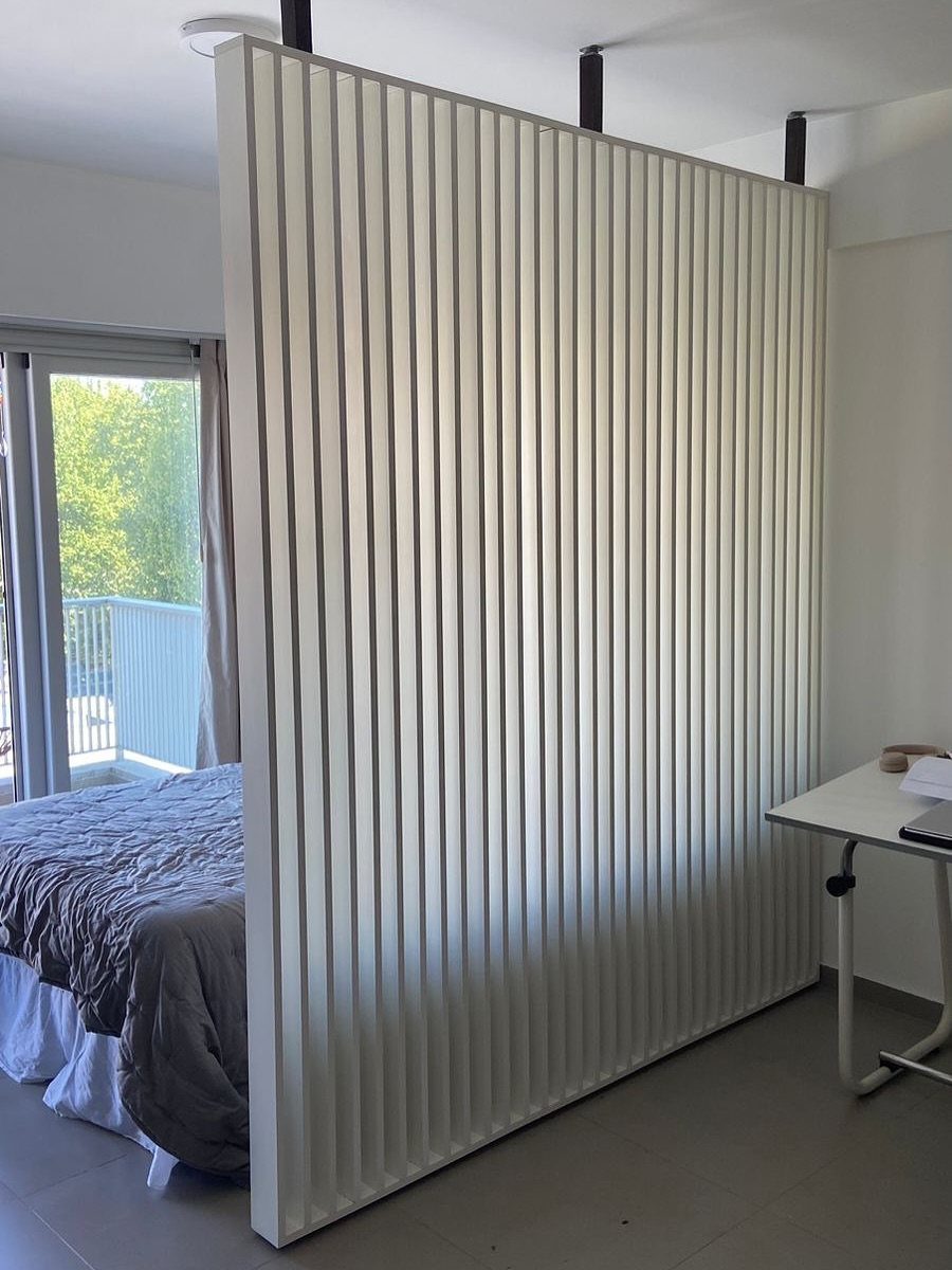 White slatted room divider separating a bed and desk in a modern studio.