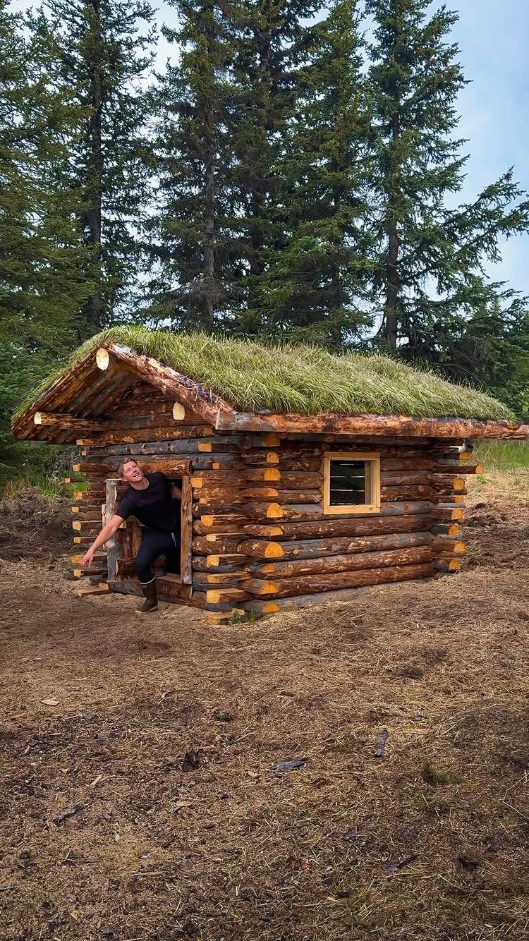 Small log cabin with a sod roof in a forest with a person exiting.