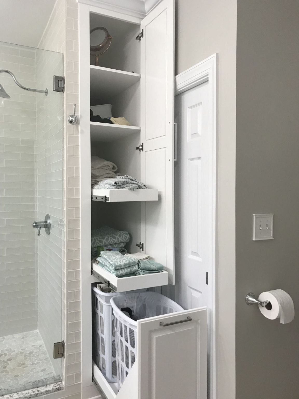 White built-in bathroom storage cabinet with shelves, pull-out drawers, and a laundry hamper.