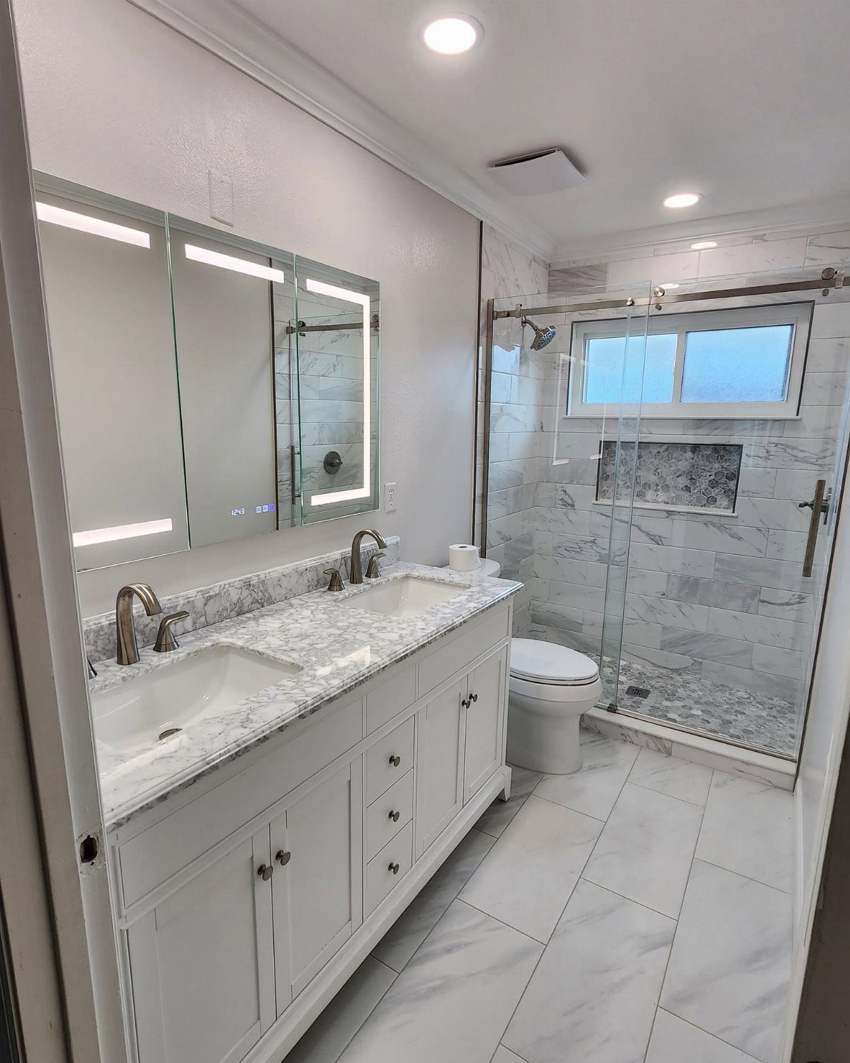 White marble bathroom with double vanity, brushed metal faucets, and glass shower enclosure.