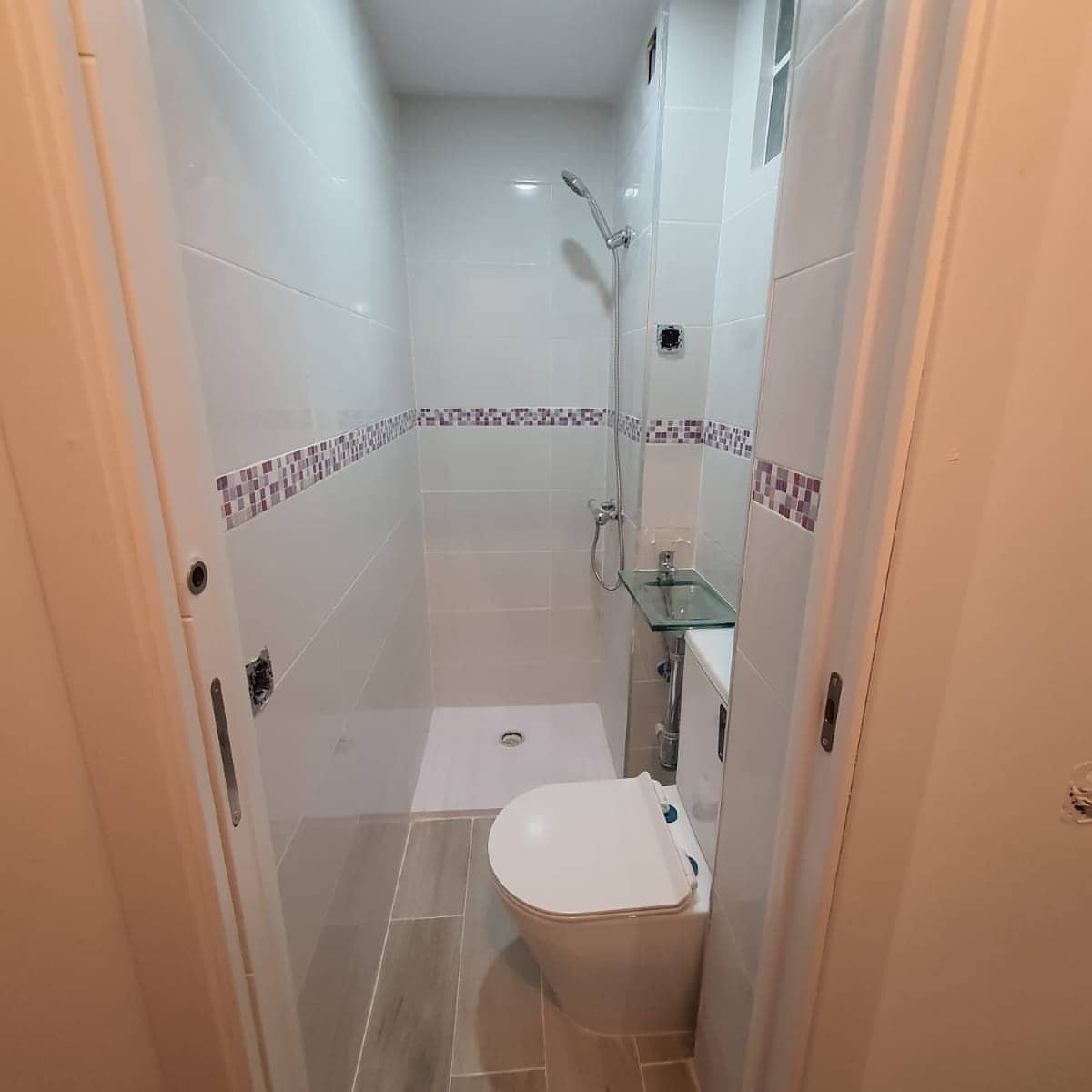 Small narrow bathroom with white tiles, purple mosaic border, and grey flooring.