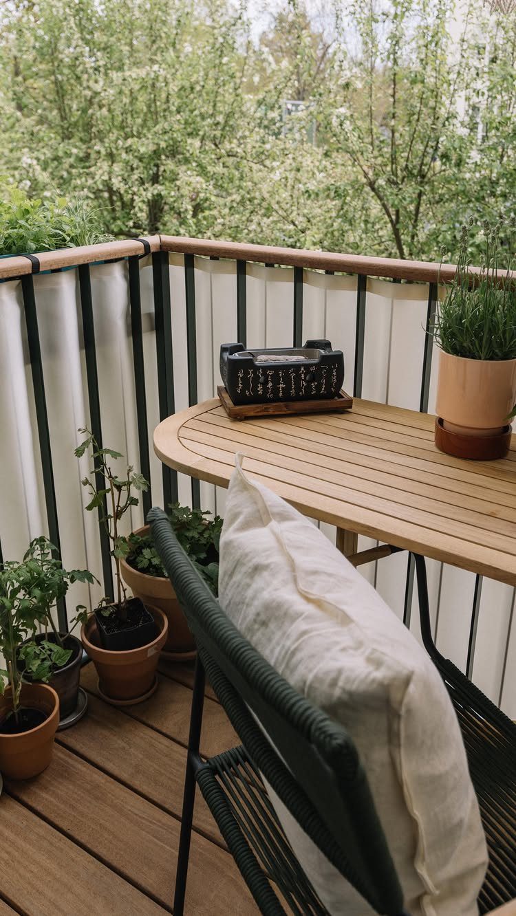 Small balcony with wooden table, black chair, white cushion, and potted plants.