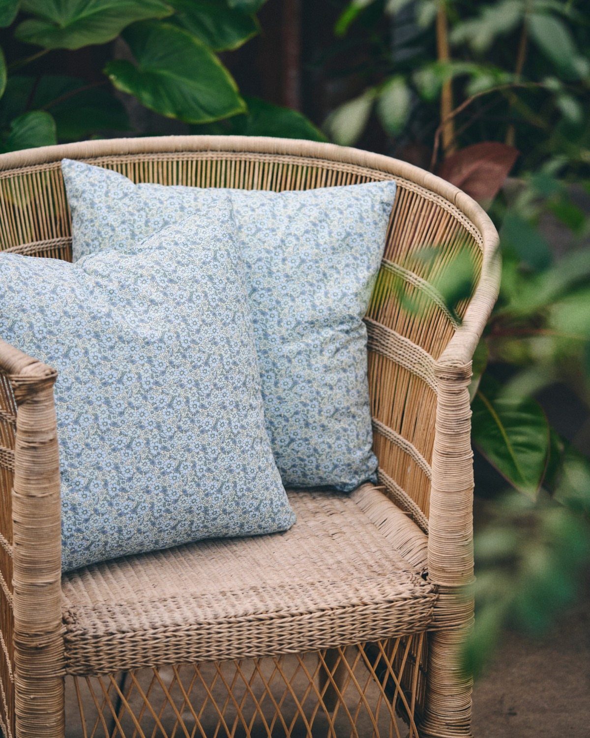 Blue floral cushions on a woven wicker chair surrounded by green tropical leaves