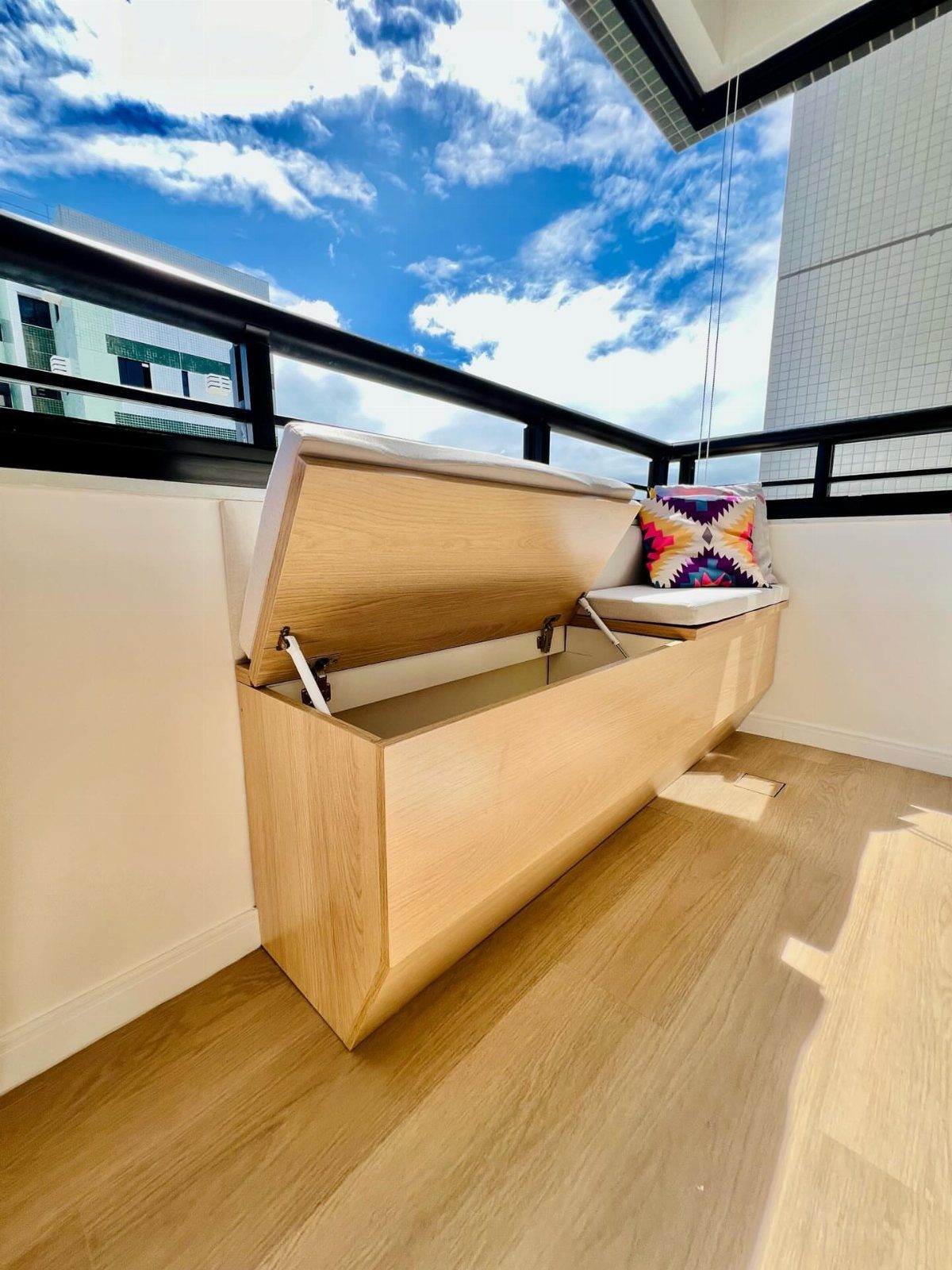 Wooden storage bench with open lid and white cushions on a sunny balcony.