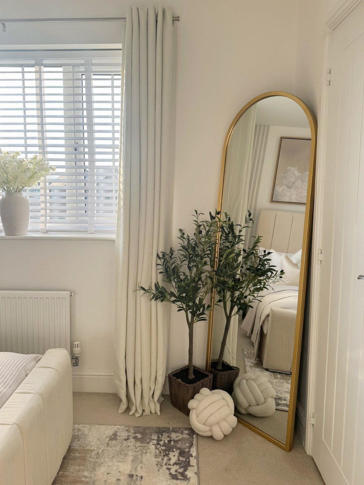 Arched gold-framed mirror reflecting a bedroom next to a window and potted plant