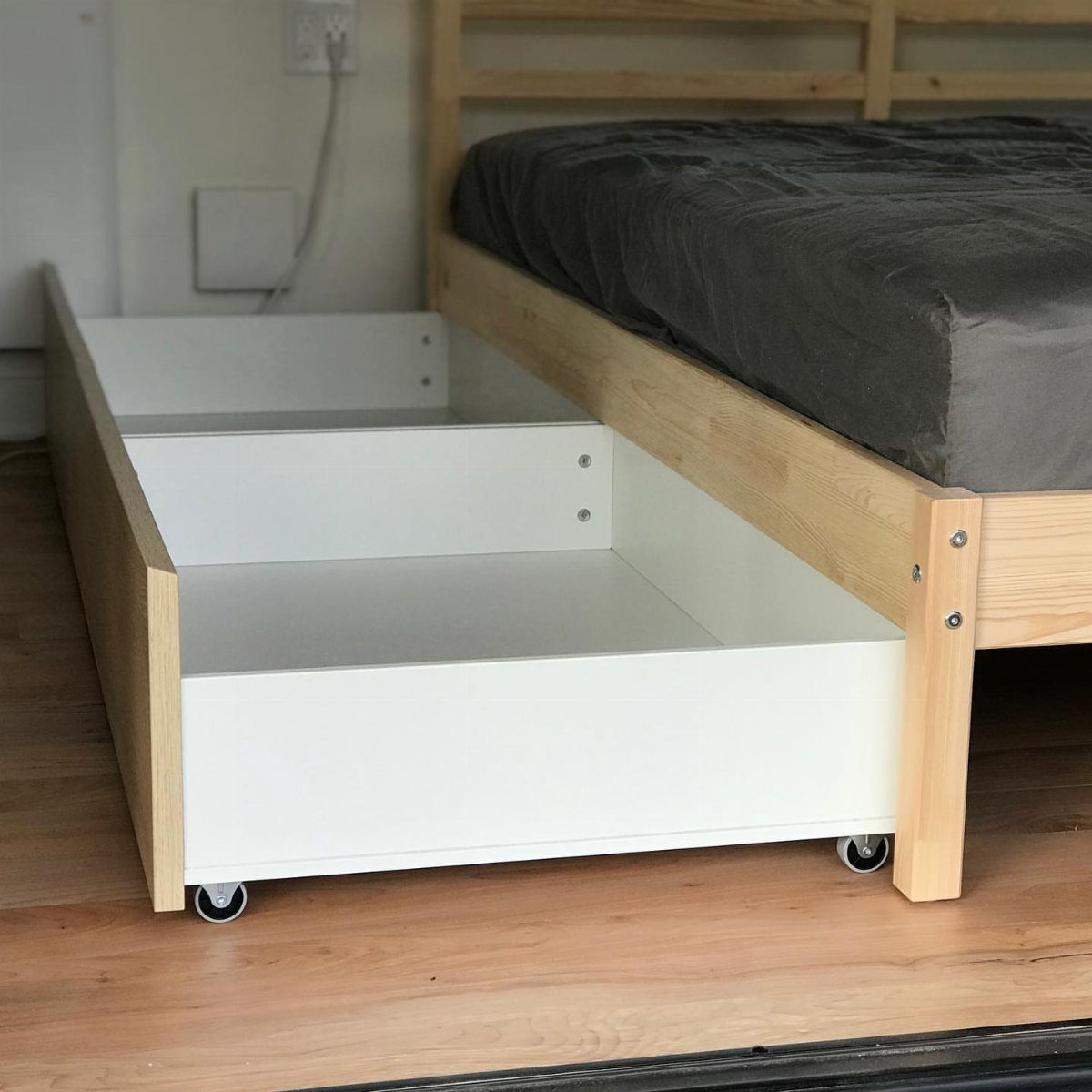 White rolling storage drawers pulled out from under a light wood bed frame.