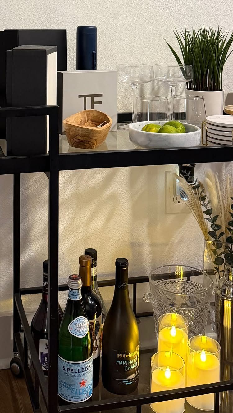 Black metal bar cart with glass shelves holding wine bottles, glasses, and candles.