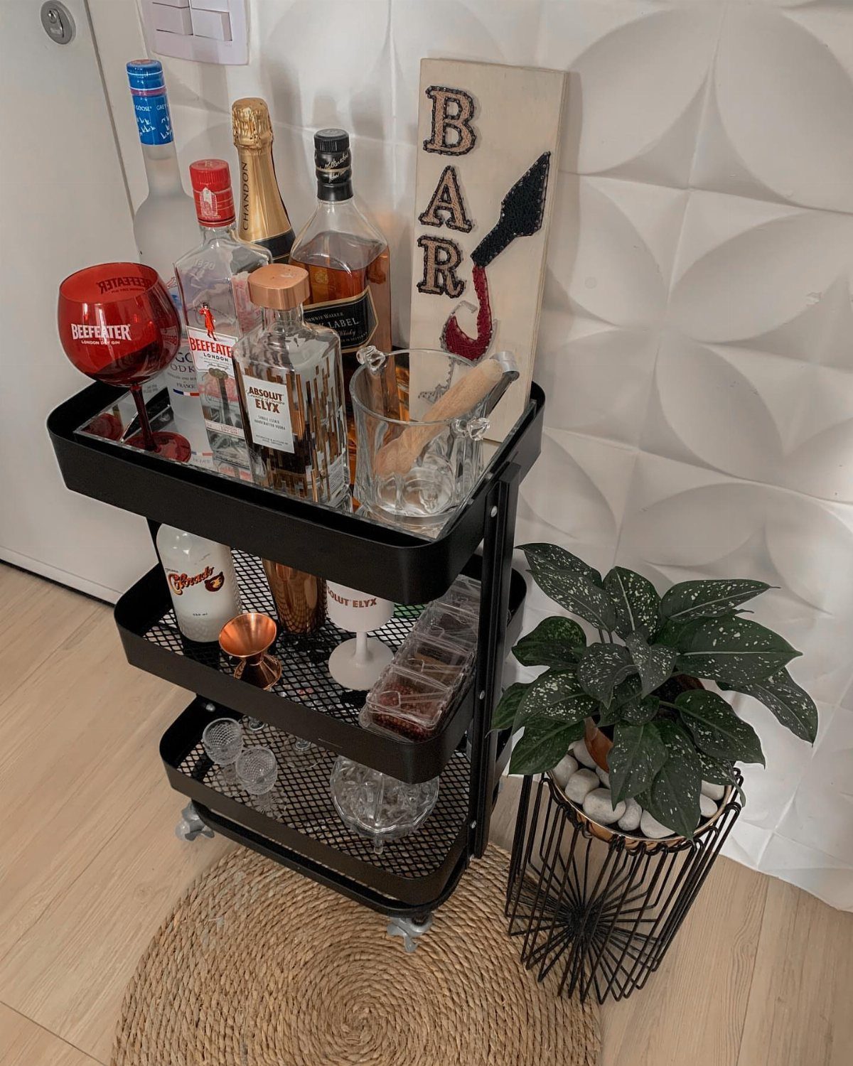 Black rolling bar cart stocked with bottles and glasses next to a potted plant.
