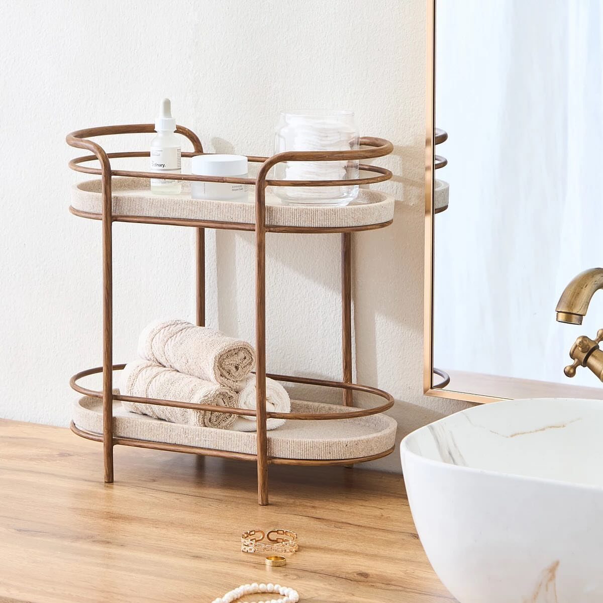 Two-tiered gold metal countertop organizer tray holding skincare products and rolled towels.