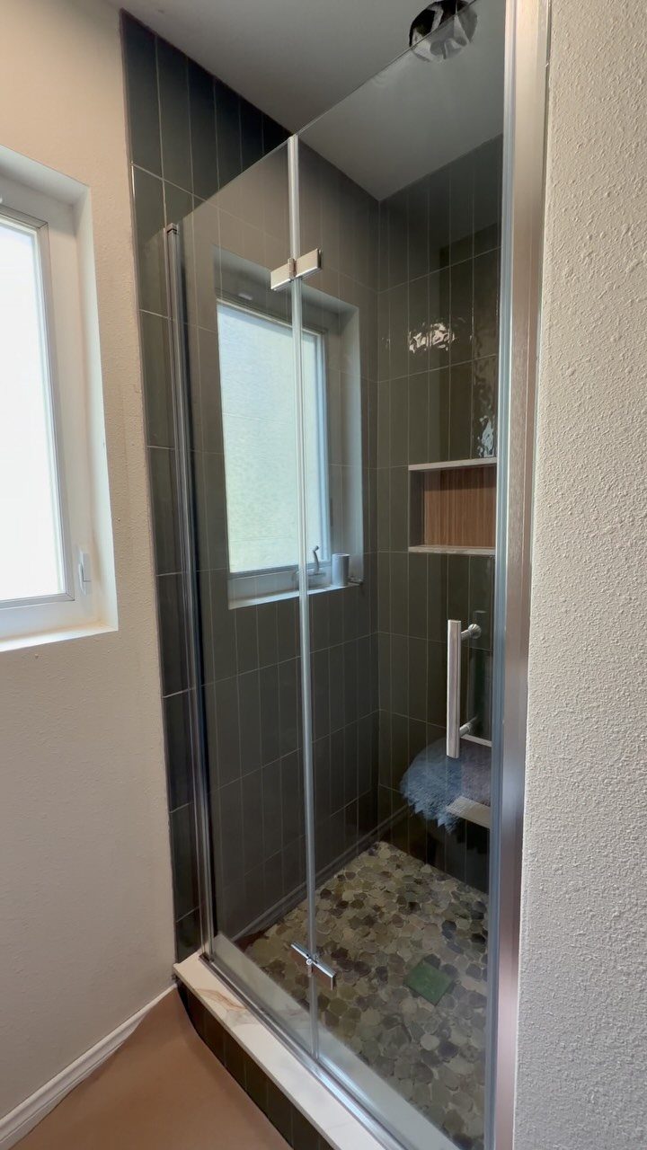 Glass shower doors with silver frames enclosing a dark tiled shower with pebble flooring.