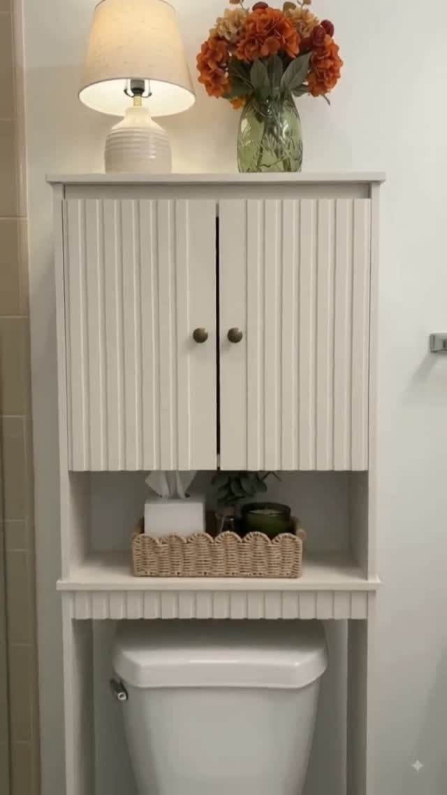 White slatted storage cabinet installed over a toilet with a lamp and flowers.