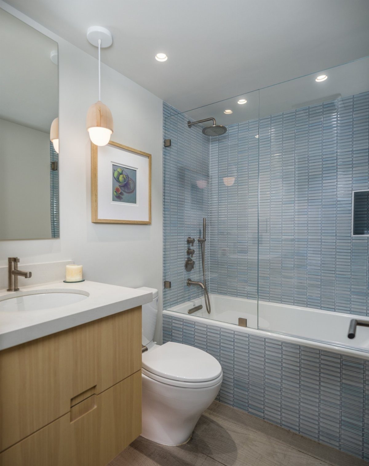 Small modern bathroom with blue tiled shower tub, white toilet, and wood vanity.