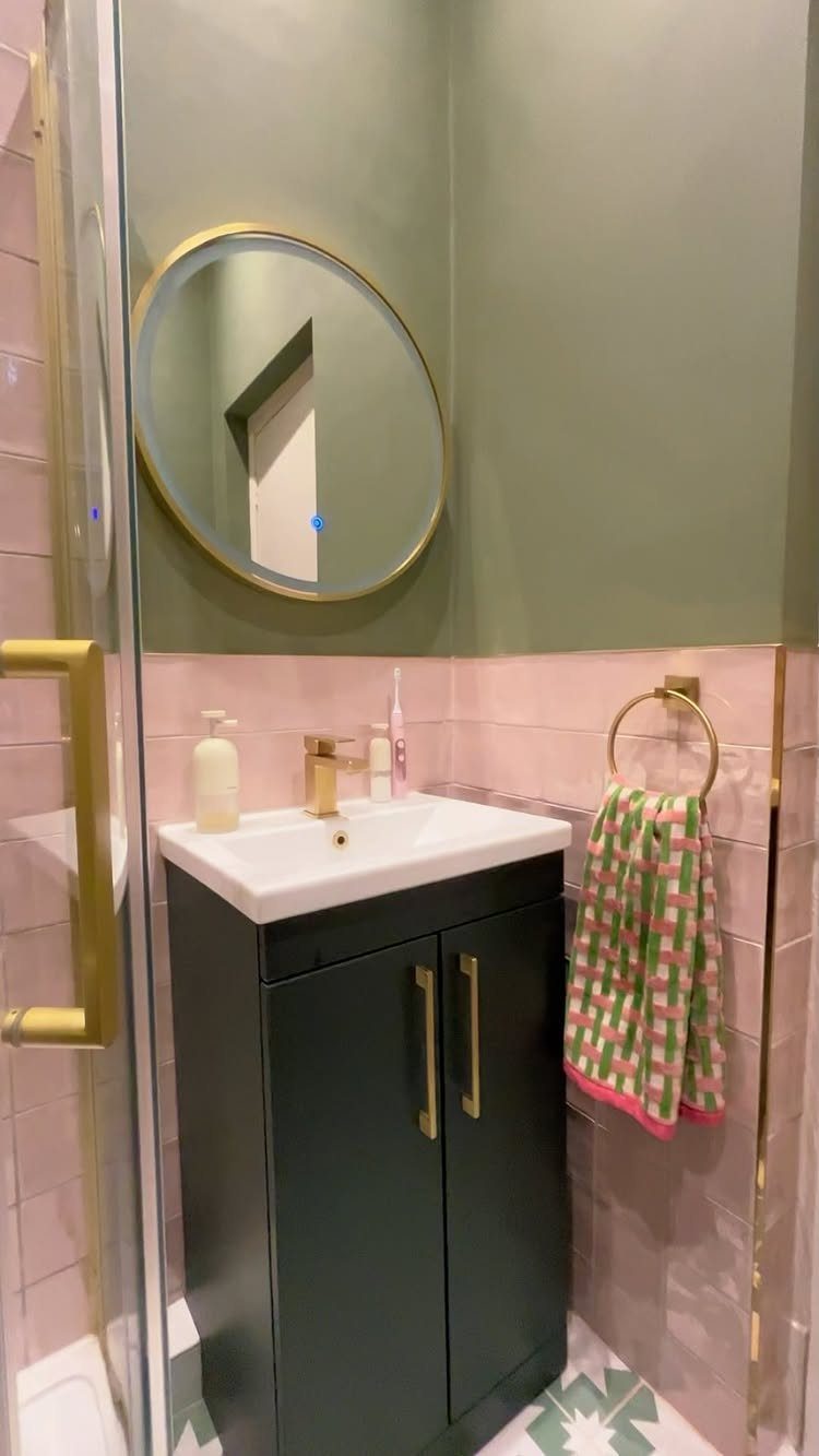 Pink tiled bathroom wall with a dark green vanity and round gold mirror.
