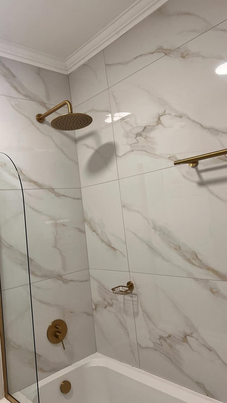 White bathtub with gold rain shower head and marble wall tiles.