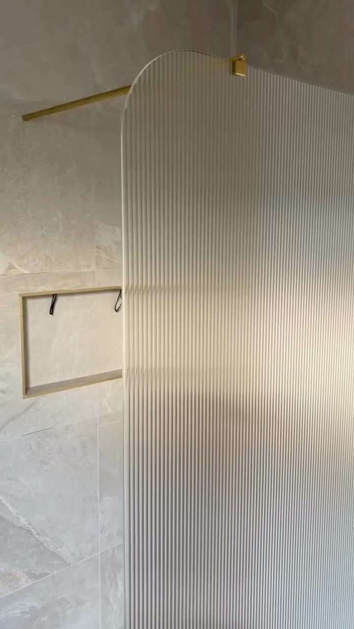Fluted glass shower panel with gold hardware against light grey marble tile walls.