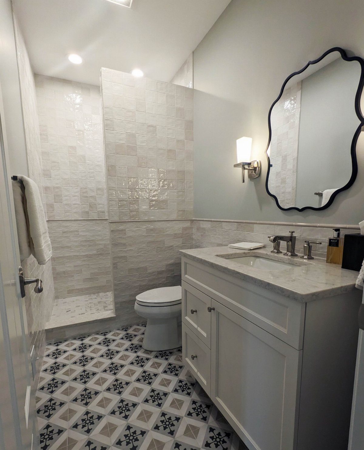 Walk-in doorless shower in a small bathroom with patterned floor tiles and white vanity.