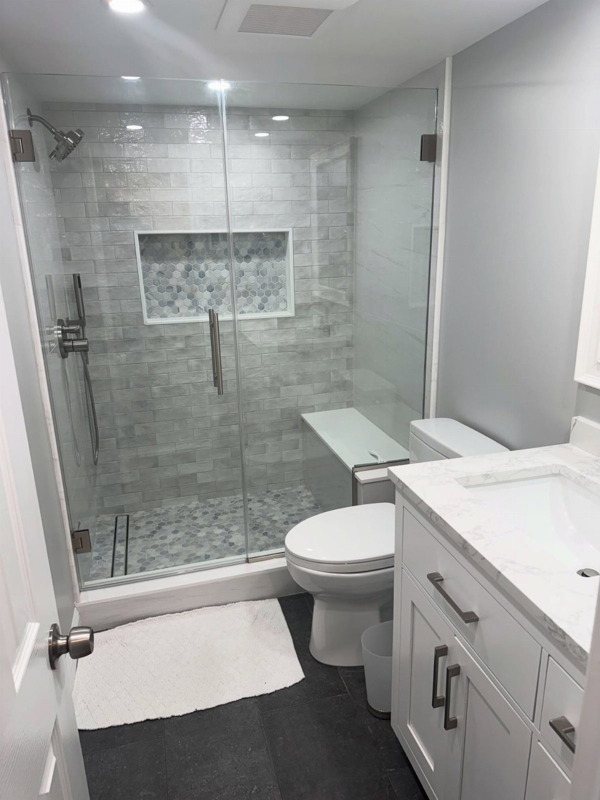 Glass shower enclosure with grey tiles, a built-in white bench, and a white vanity.