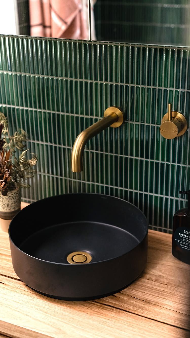Black vessel sink on wood counter with vertical green tiles and gold faucet.