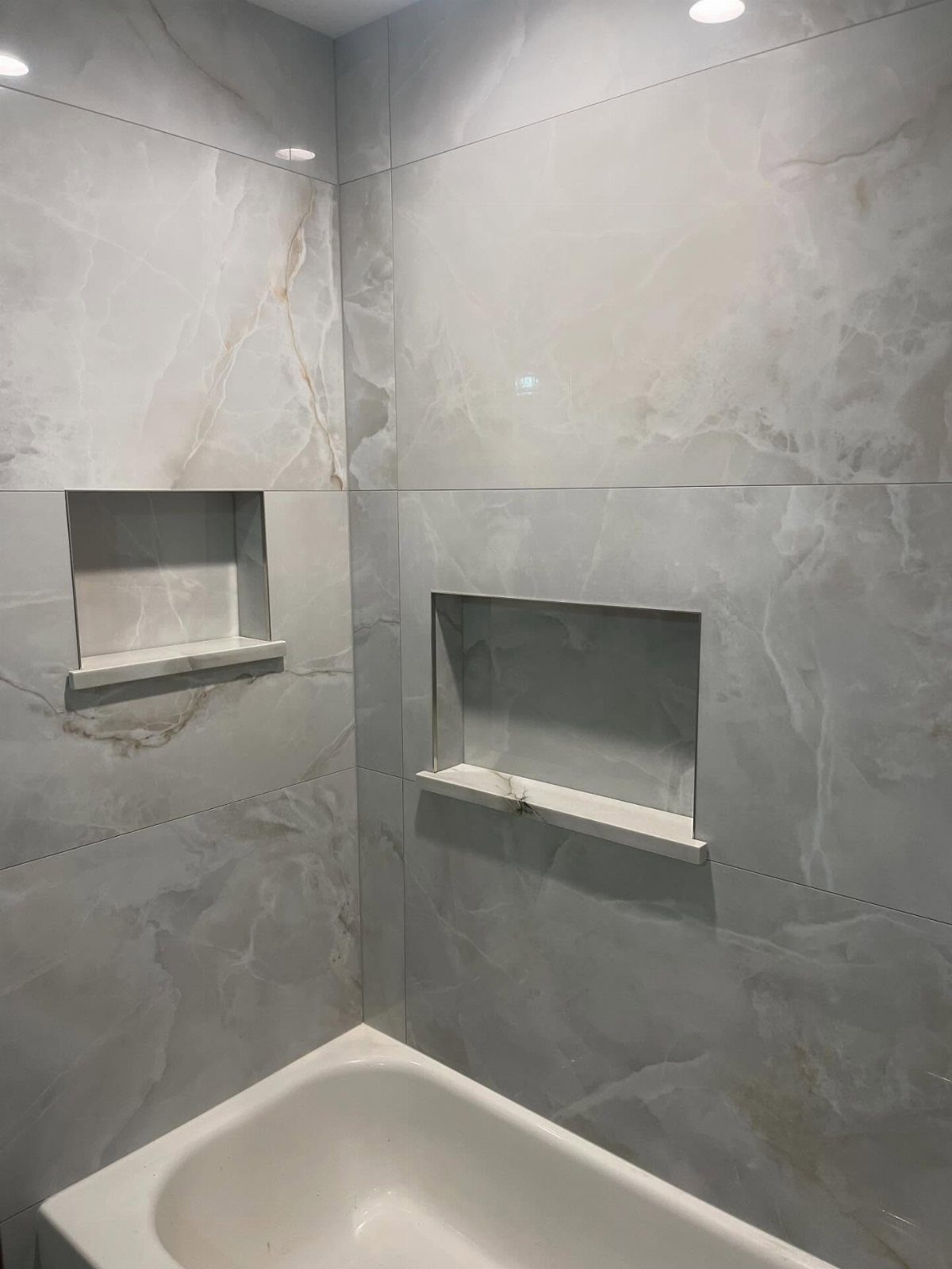 White bathtub with large grey marbled wall tiles and two built-in storage niches.