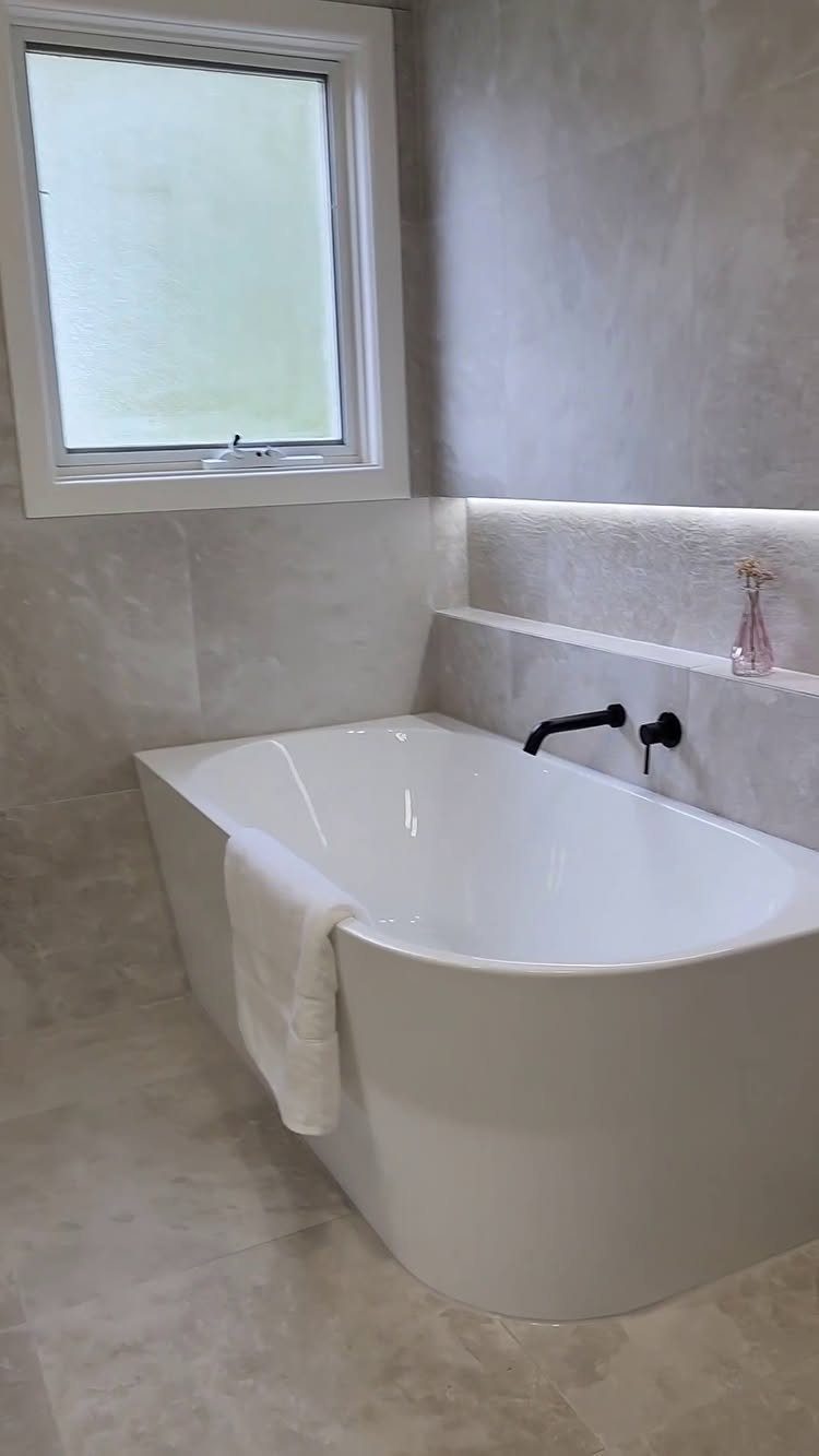 White freestanding bathtub in a grey stone bathroom next to a window