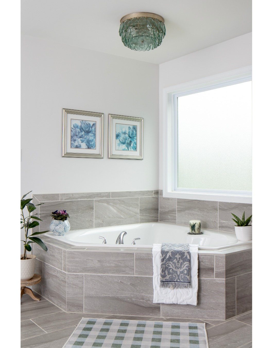 White corner bathtub with grey tile surround next to a frosted window