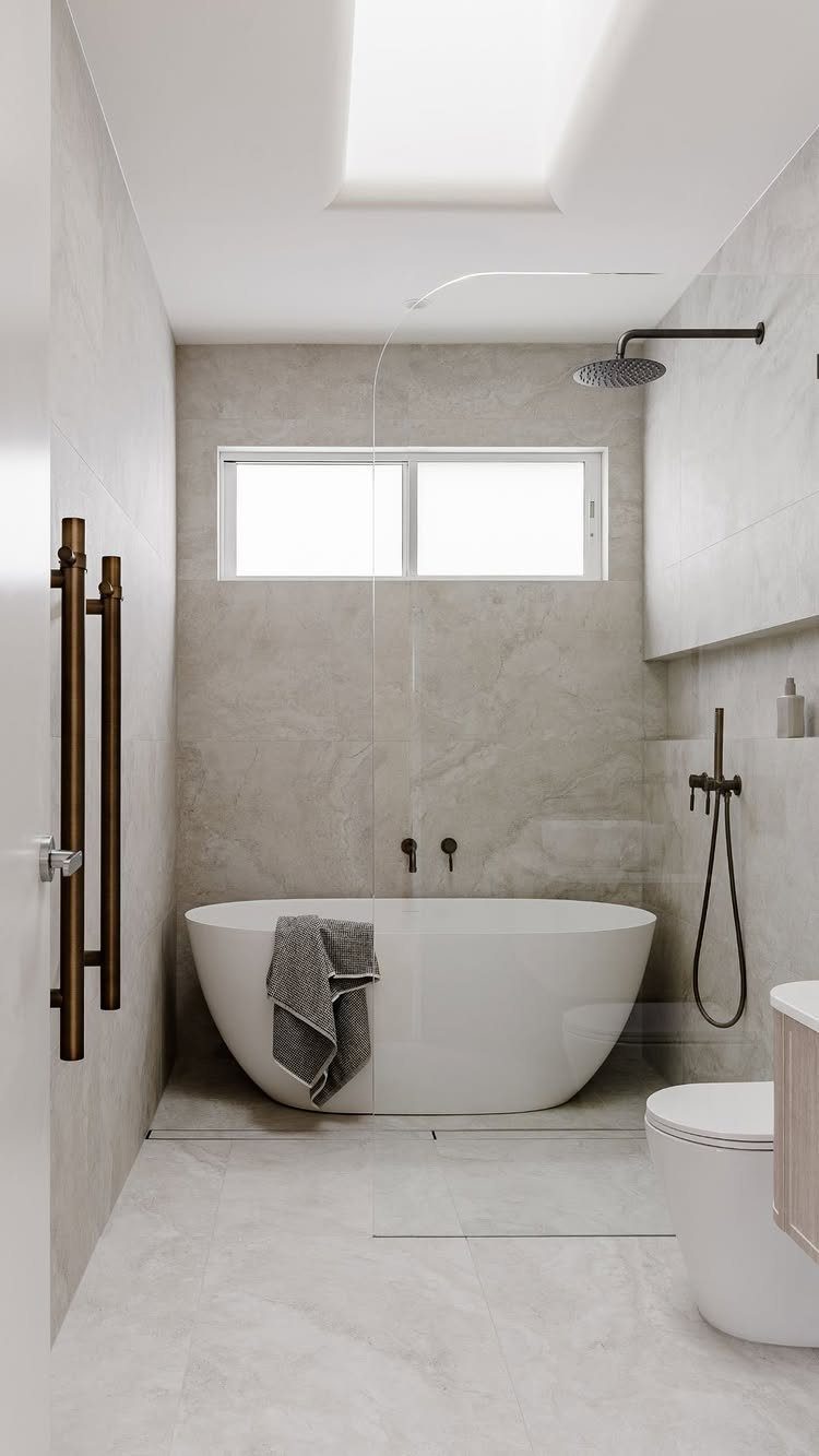 Modern bathroom with large grey marble tiles, white freestanding tub, and glass shower panel.