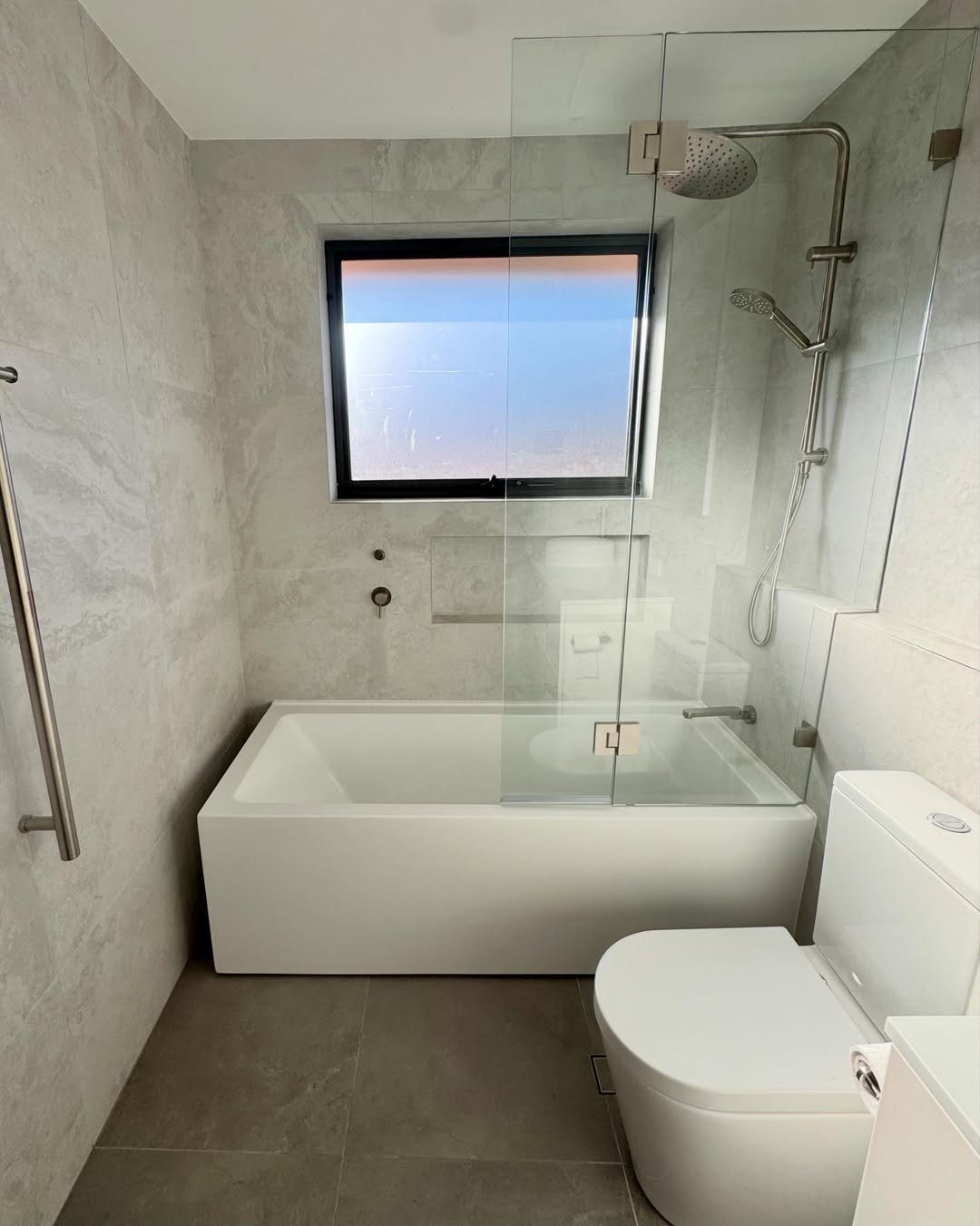 White rectangular bathtub positioned directly beneath a black-framed window in a modern bathroom.