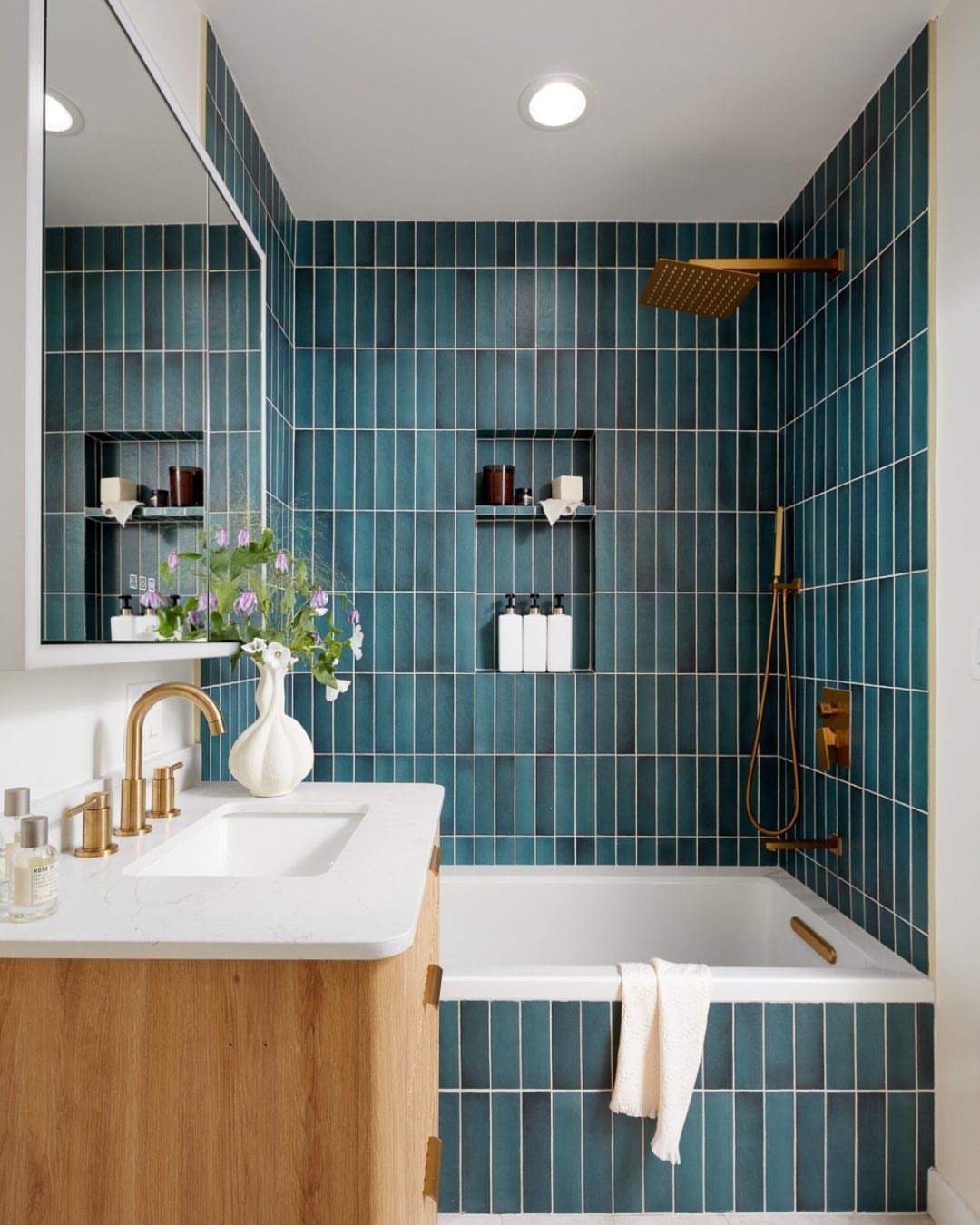 Teal vertical tiled bathroom with a white bathtub and gold fixtures.