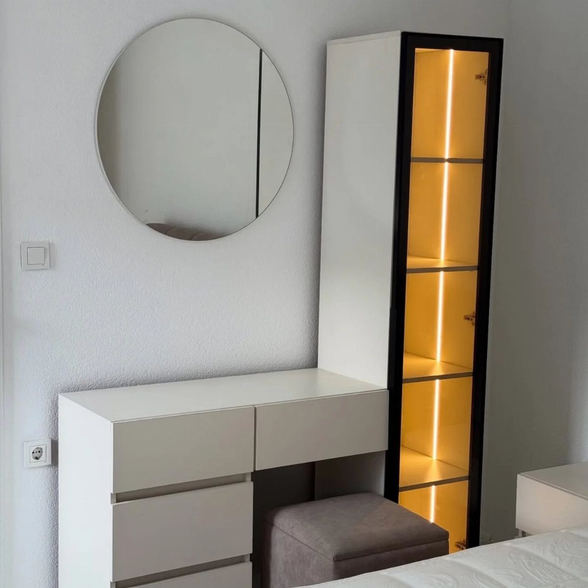 White vanity desk and illuminated tall cabinet with a round wall mirror.