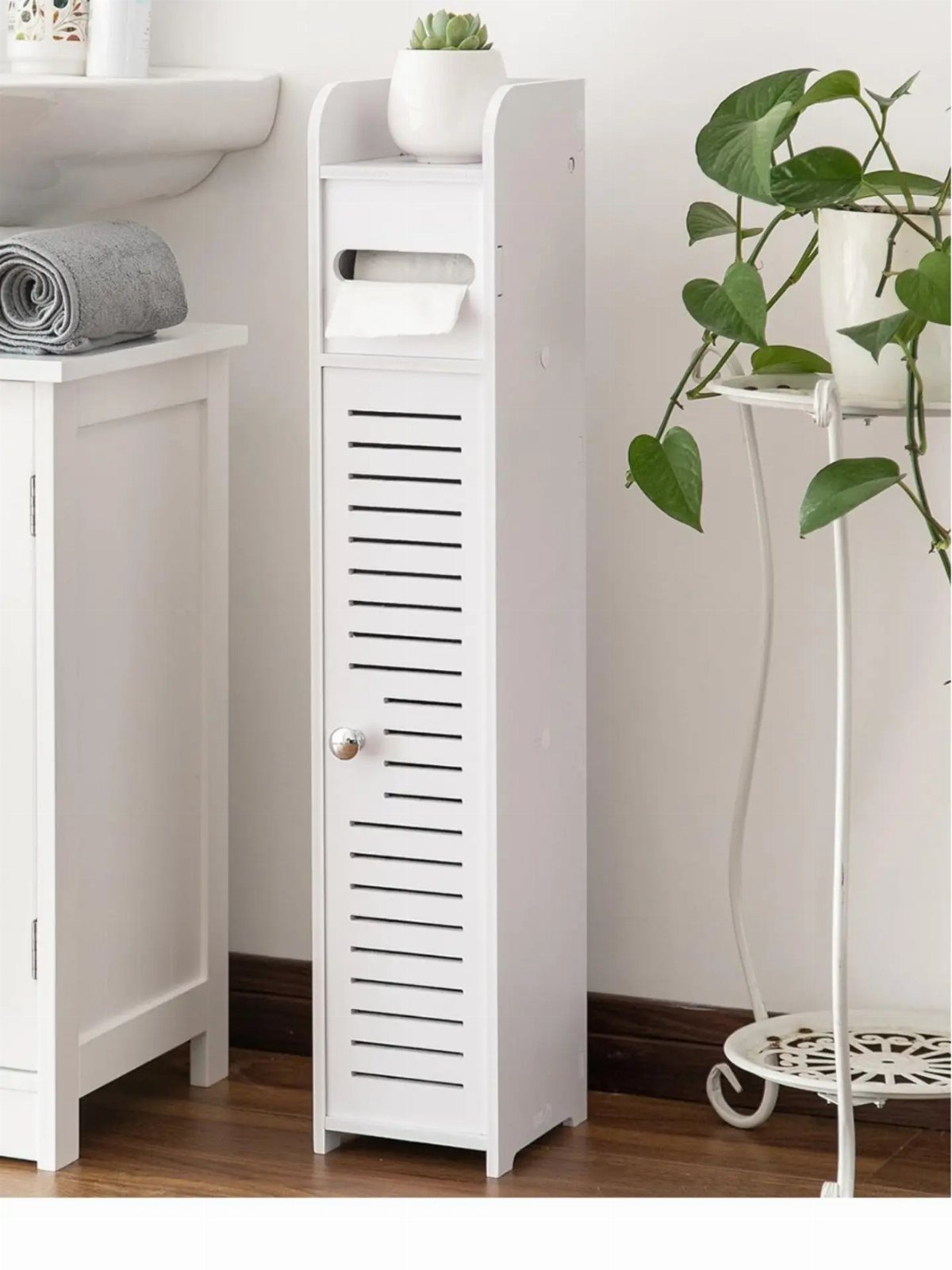 Slim white bathroom storage cabinet with a slatted door and small potted plant.