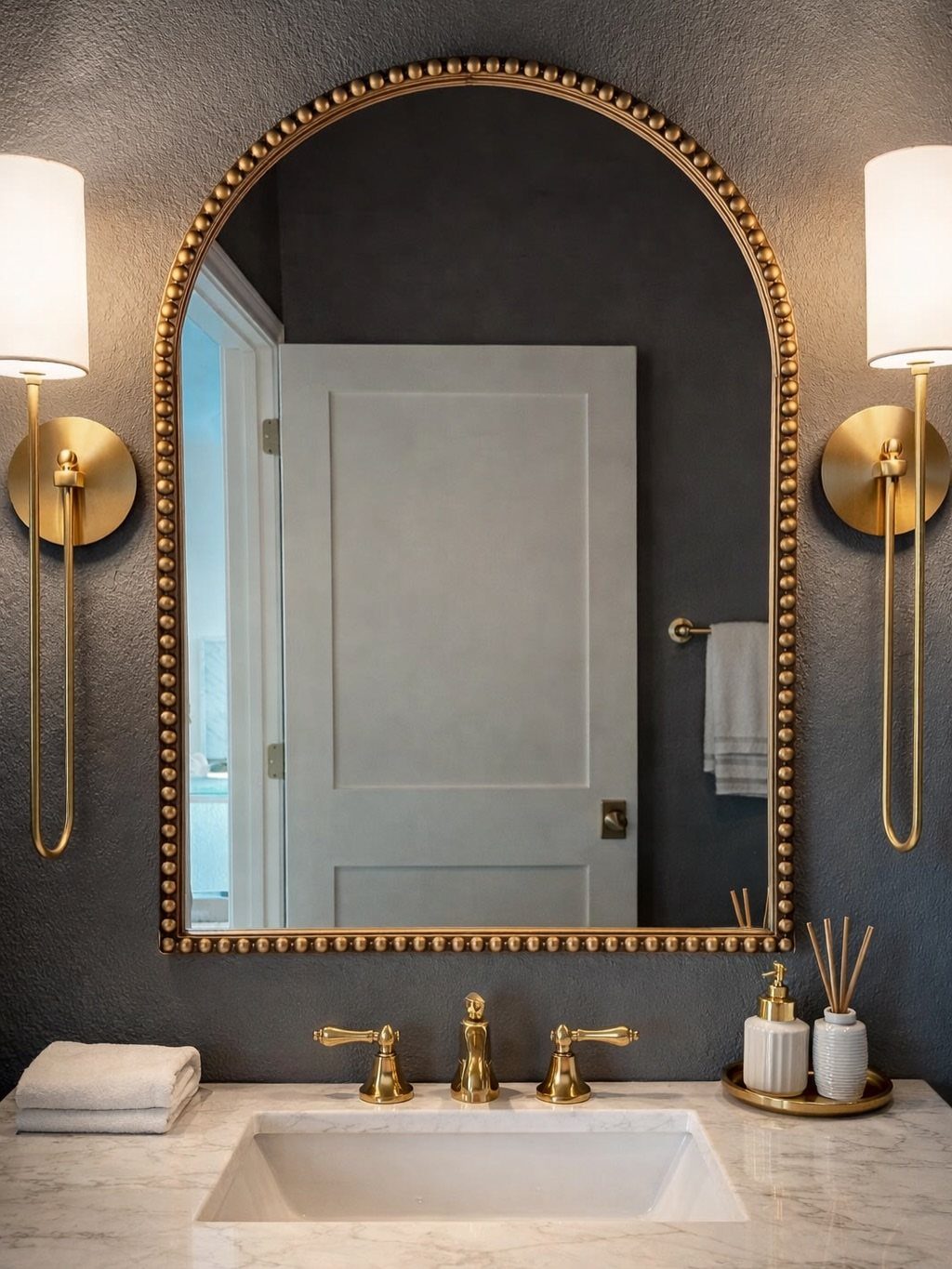 Arched gold beaded mirror centered between two gold sconces above a marble sink