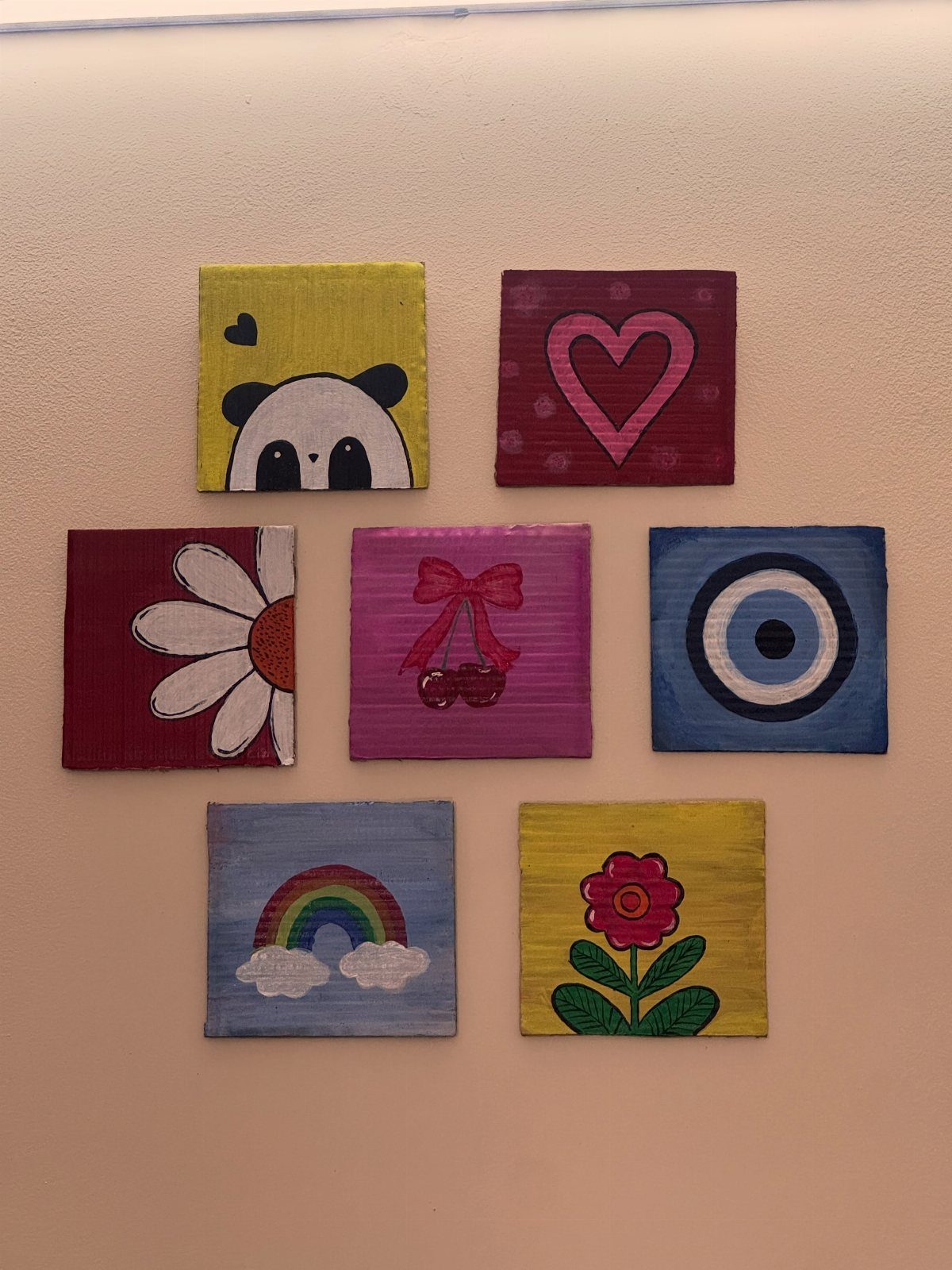 Seven small square colorful paintings arranged in a grid on a white wall.