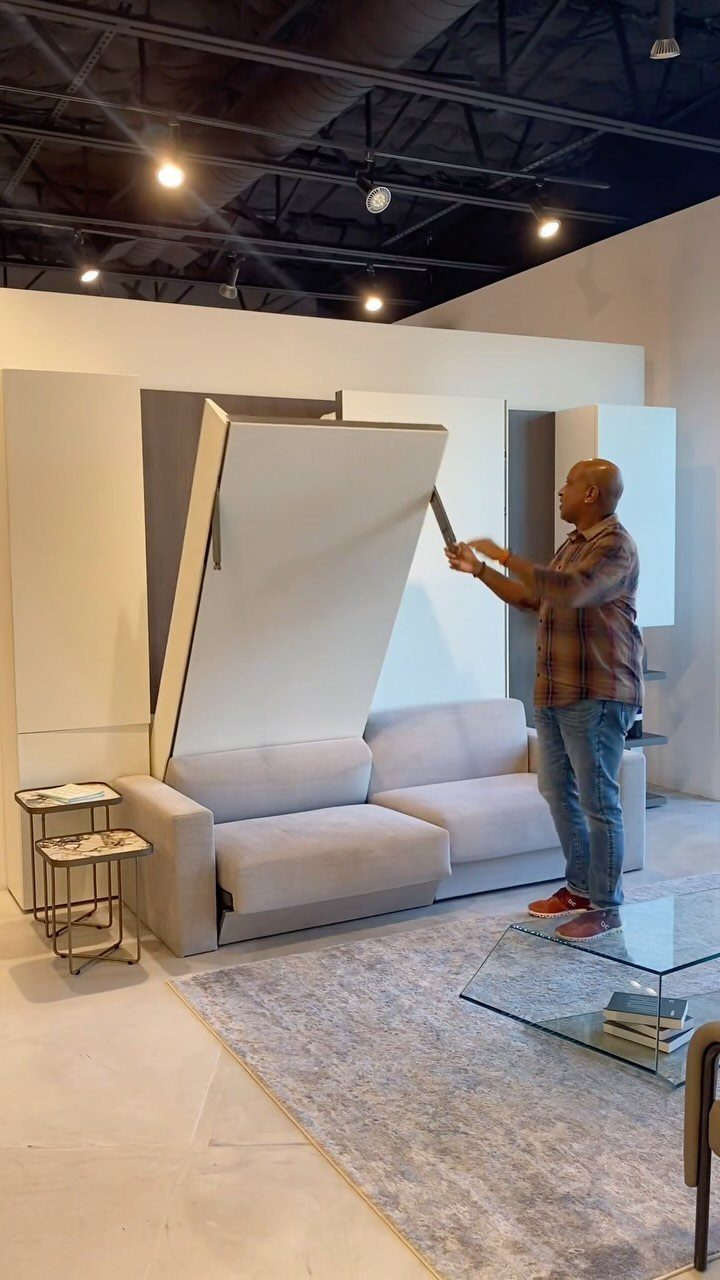 Man pulling down a white Murphy bed above a grey sofa in a showroom.