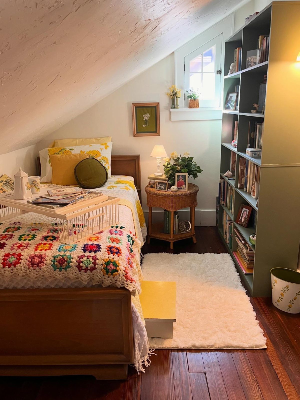 Cozy attic bedroom with sloped ceiling, colorful crochet blanket, and tall grey bookshelf.