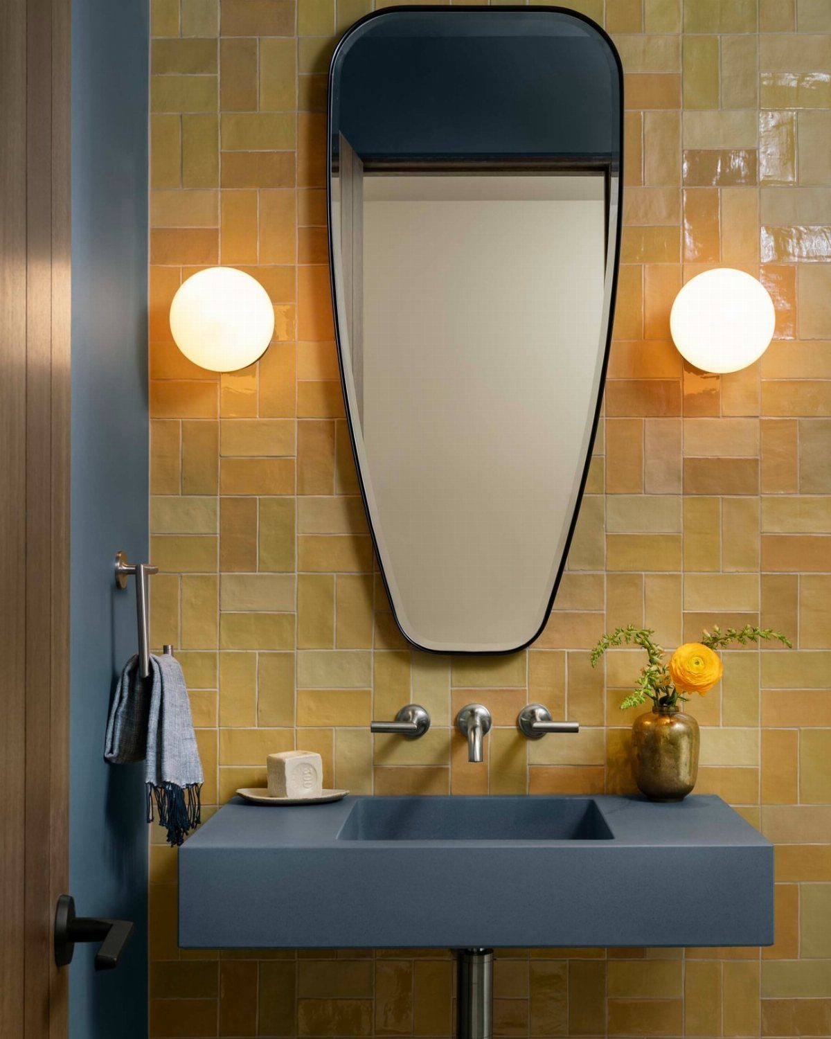 Blue rectangular sink and oval mirror against yellow tiled walls with globe lights.