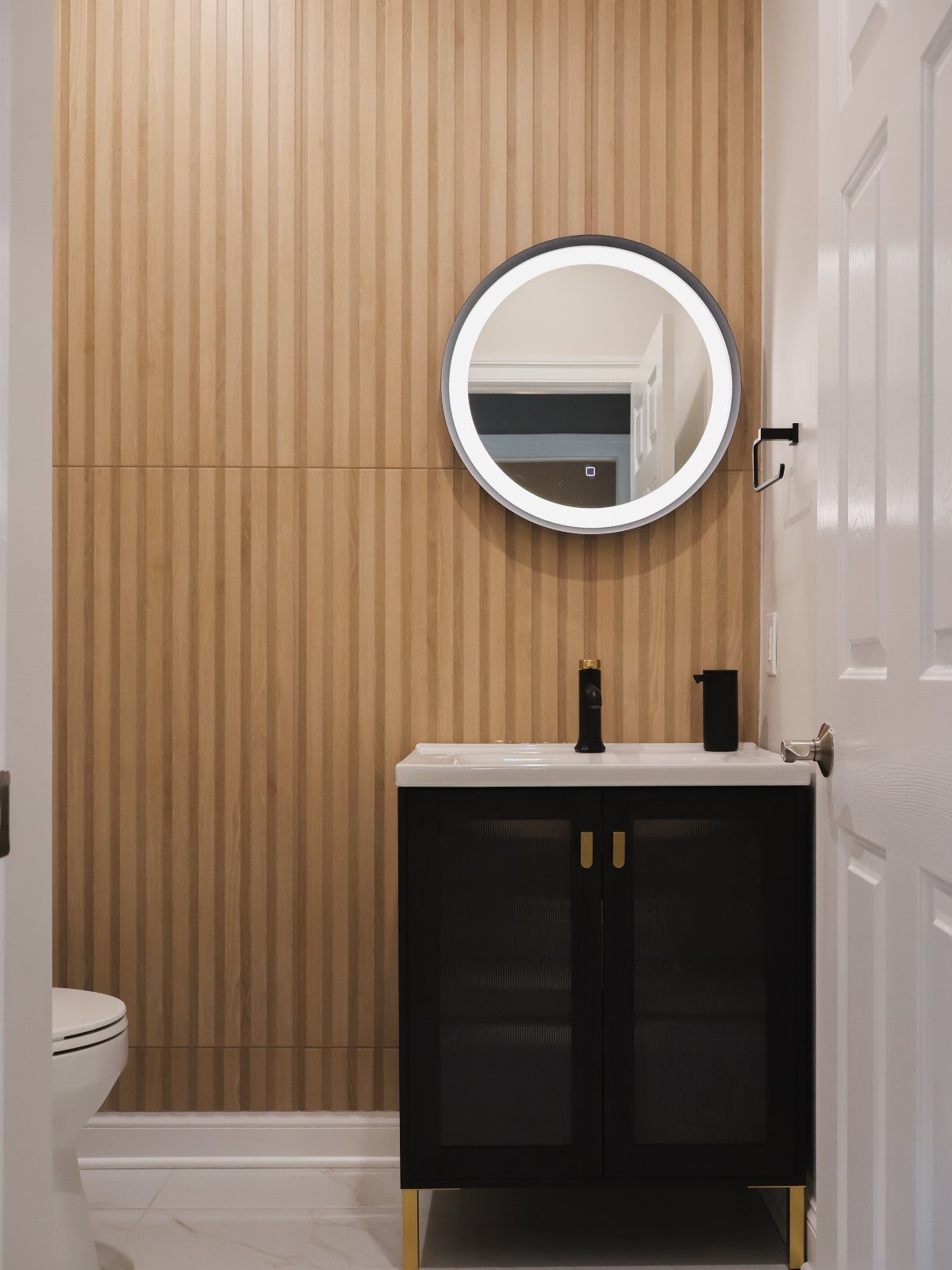 Black vanity with gold hardware against a vertical wood slat wall and round mirror.