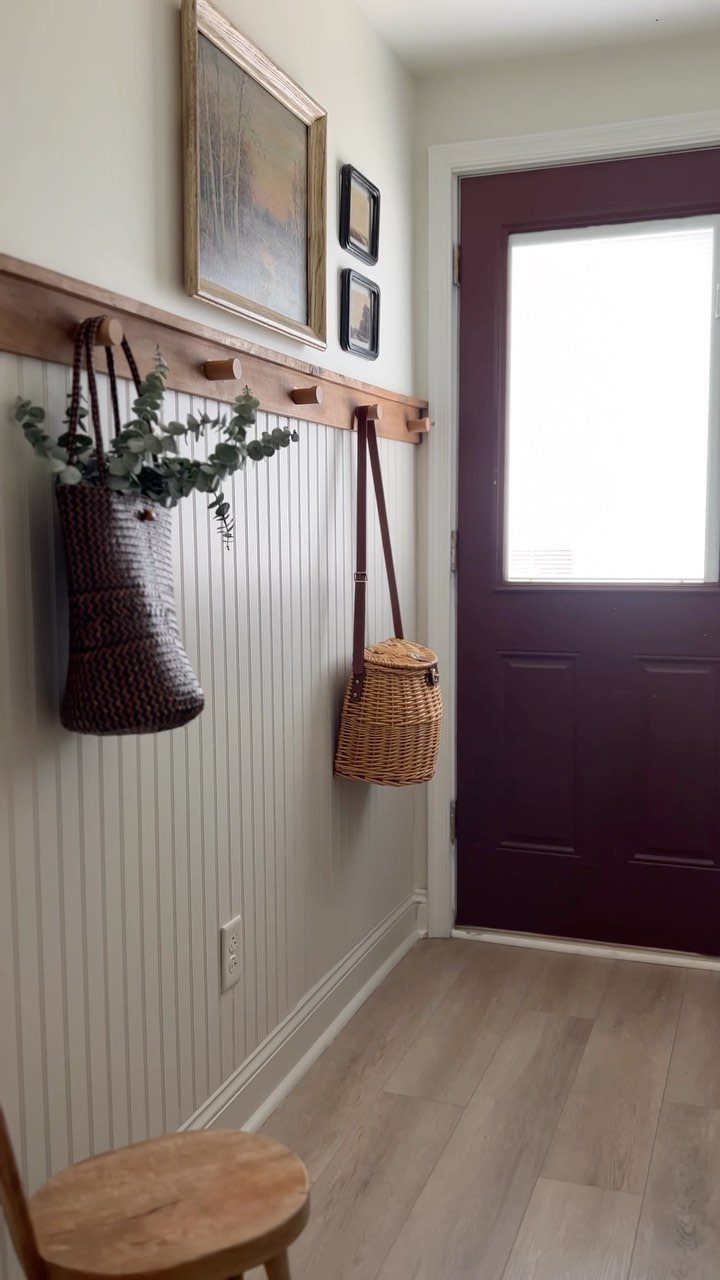 Wooden rail with hooks holding woven baskets on a white beadboard entryway wall.