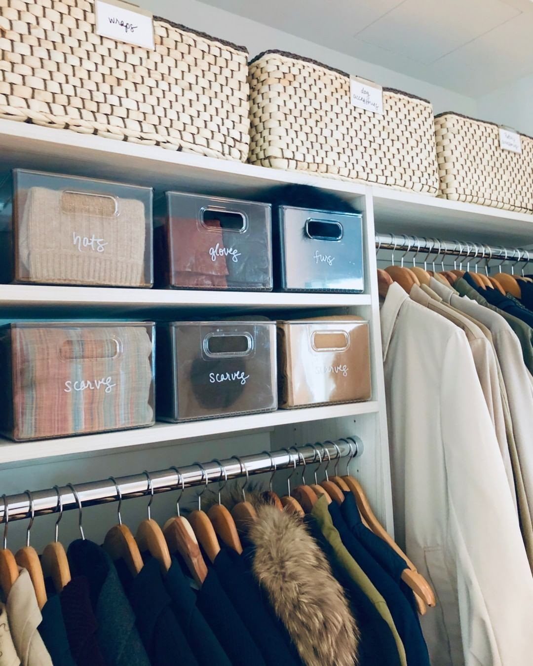 Labeled clear plastic bins and woven baskets on white closet shelving with hanging clothes.