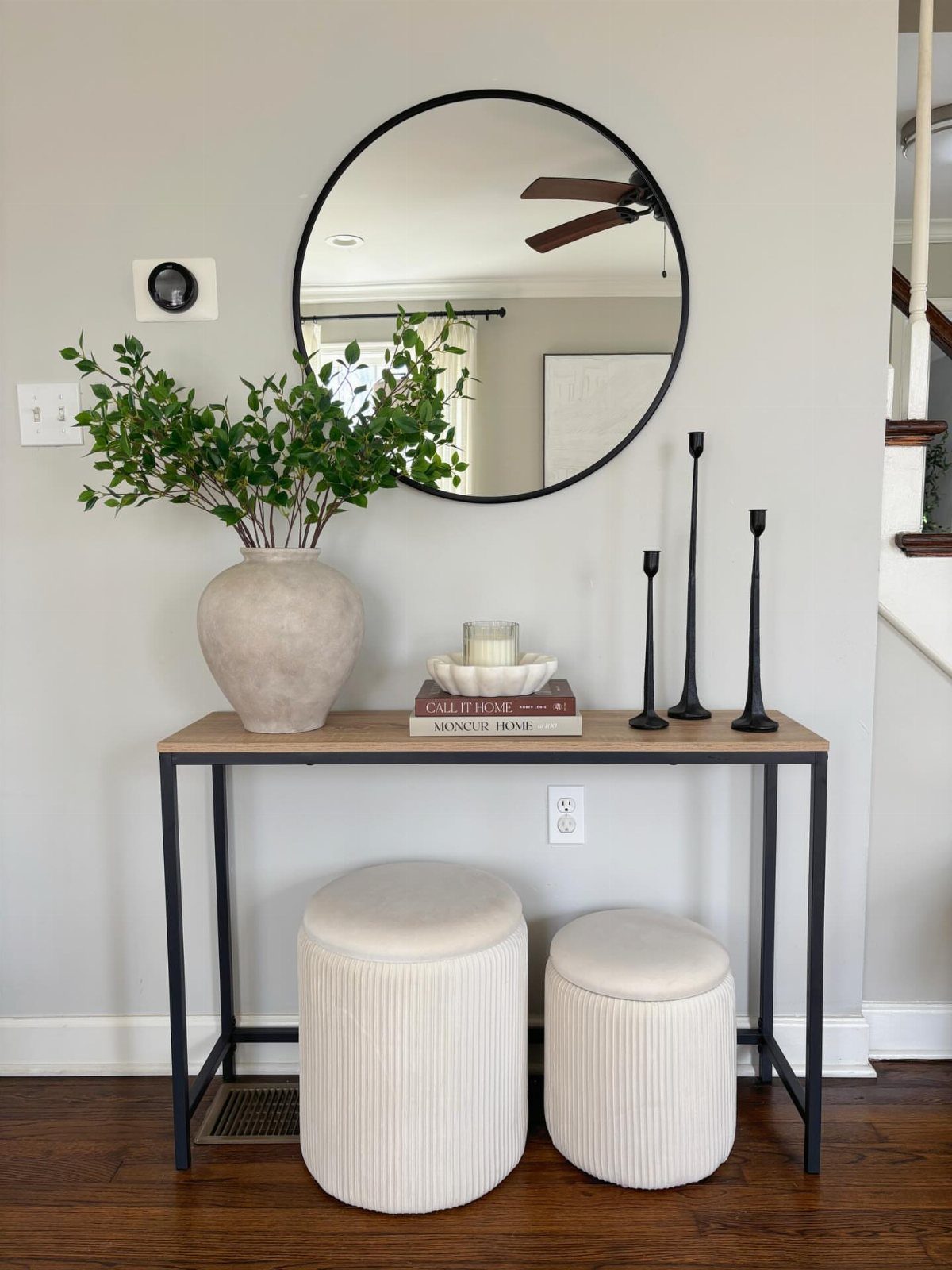 Narrow wooden console table with a round mirror and white ottomans in an entryway.