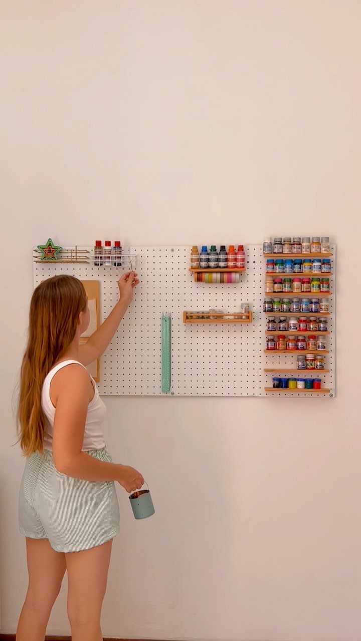 Woman organizing colorful paint bottles on a white wall-mounted pegboard storage system