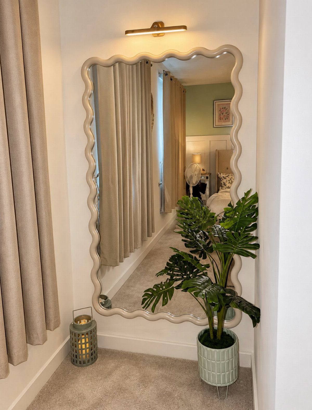Floor-length mirror with wavy frame reflecting a bedroom, next to a potted plant.