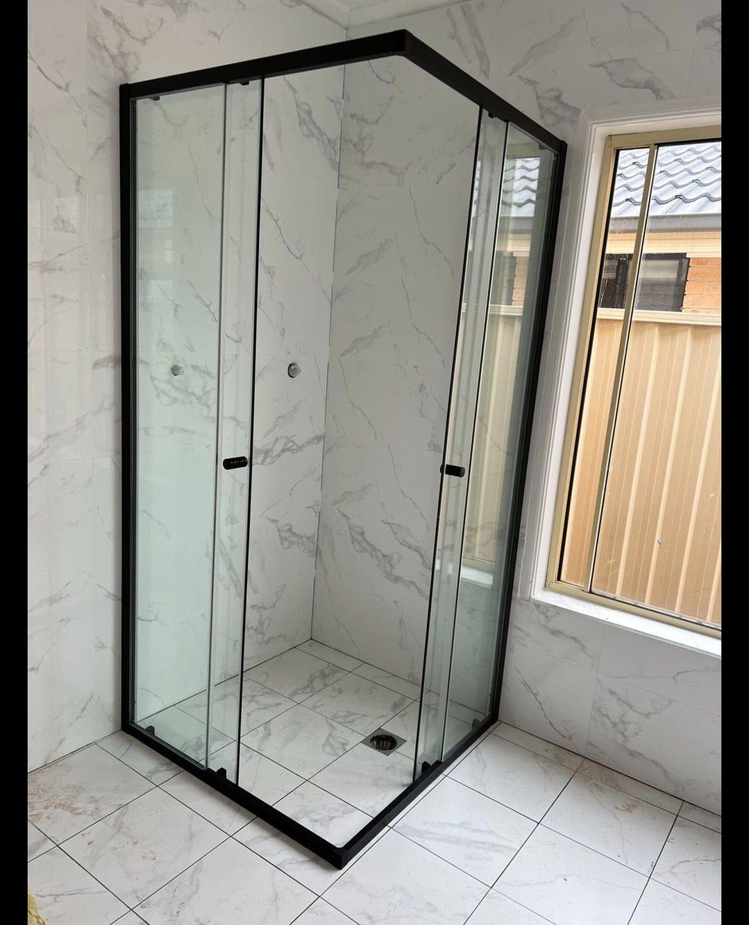 Corner walk-in shower with black frames and white marble tiles in a bright bathroom.