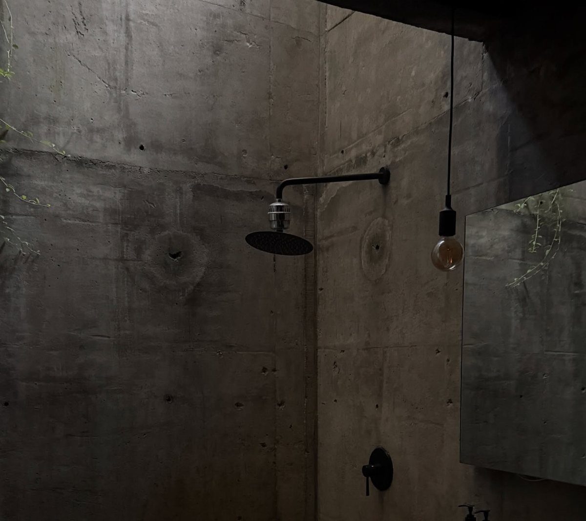 Concrete shower walls with a black metal rain showerhead and hanging light bulb.