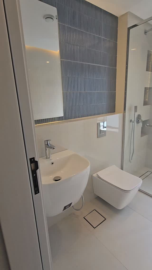 Modern small bathroom with white fixtures, grey vertical tiles, and glass shower enclosure.