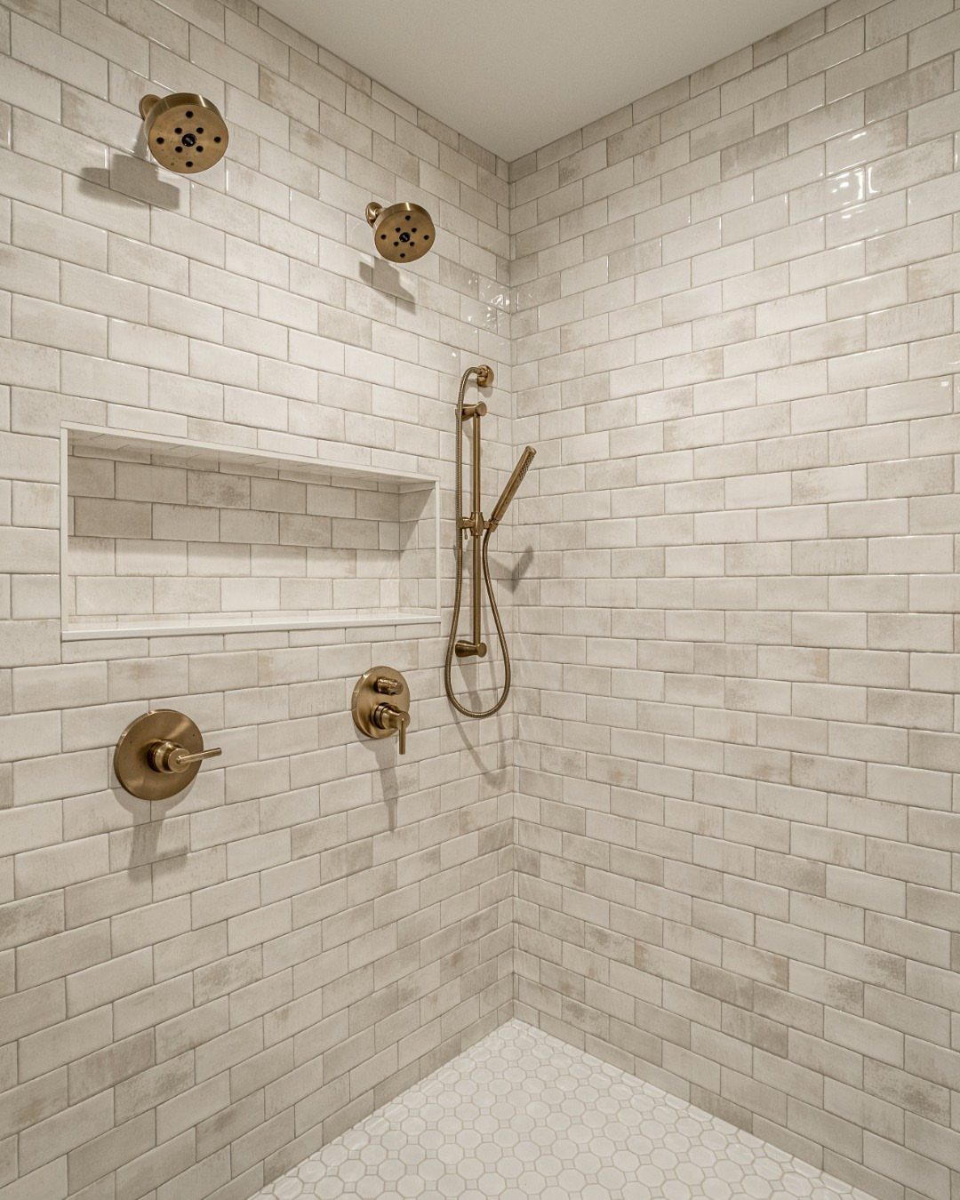 Two gold showerheads and a handheld sprayer on white subway tile walls.