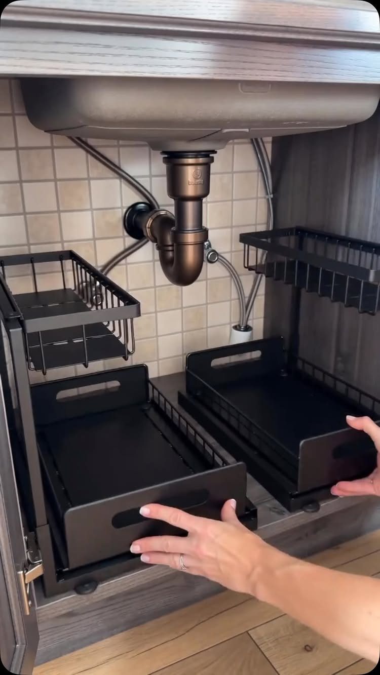 Black sliding organizers under a kitchen sink with plumbing pipes and tiled walls.