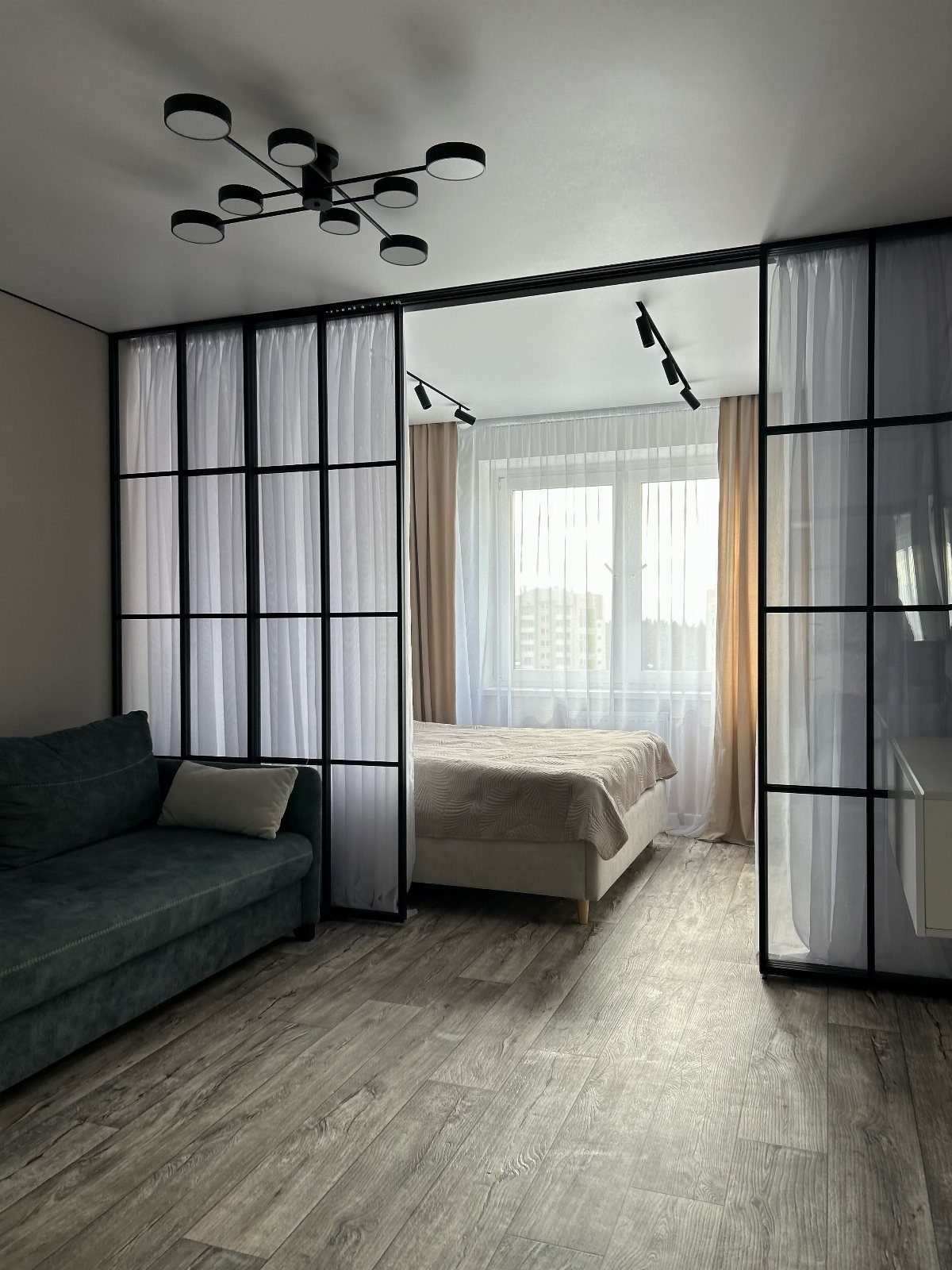 Black metal grid frame with white curtains separating a living area from a bedroom