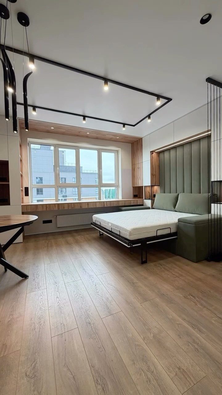 Modern studio apartment interior with light wood floors, green bed, and track lighting.