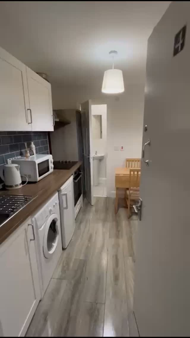 Narrow studio apartment interior with white kitchenette and small wooden dining table.
