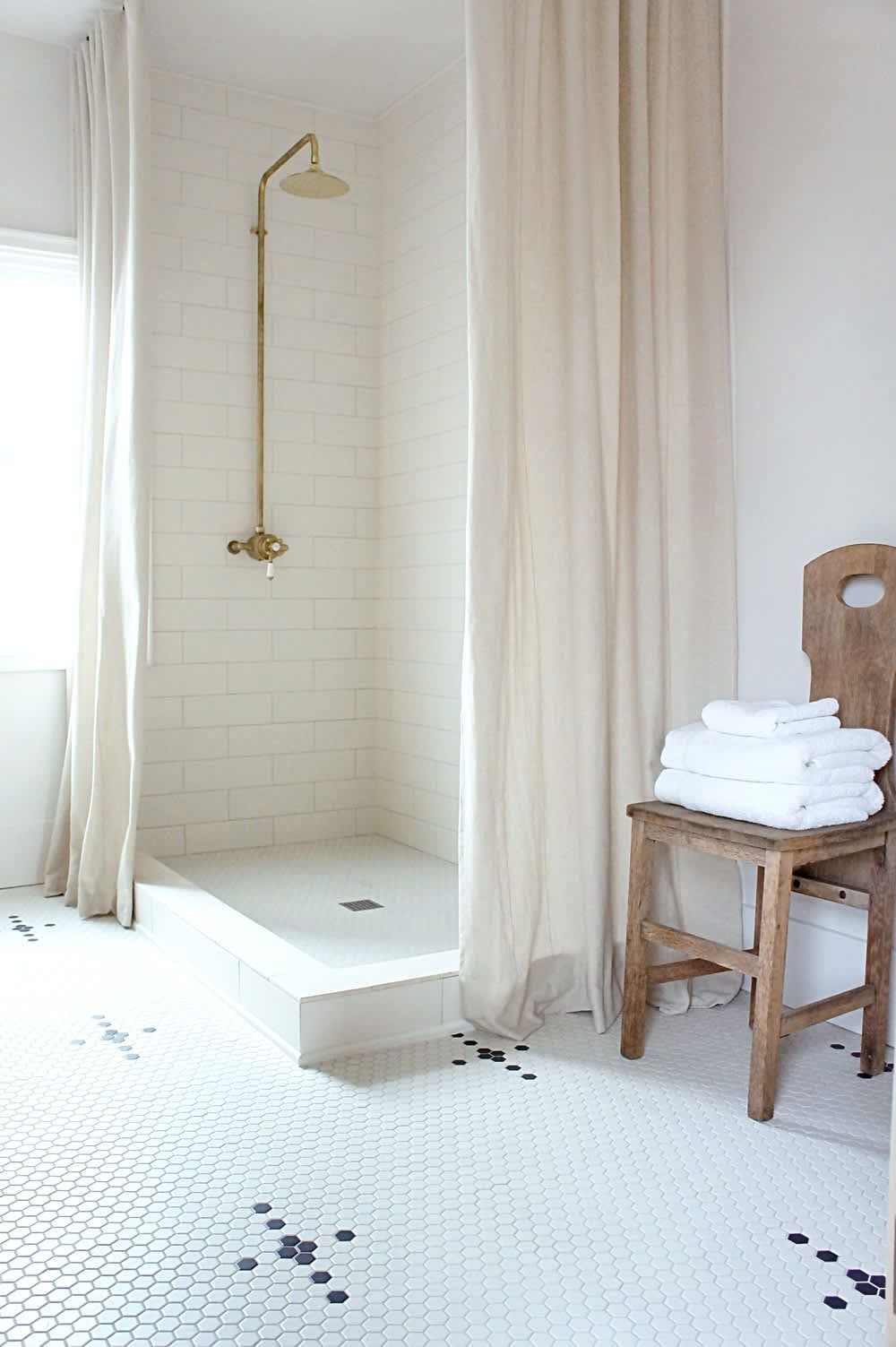 Walk-in shower with cream curtains, gold fixtures, and white hexagonal tiled flooring.